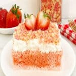 Strawberry crunch tres leches cake on a white plate. Next to a jar filled with strawberry shortcake crumble topping and a bowl of fresh strawberries.