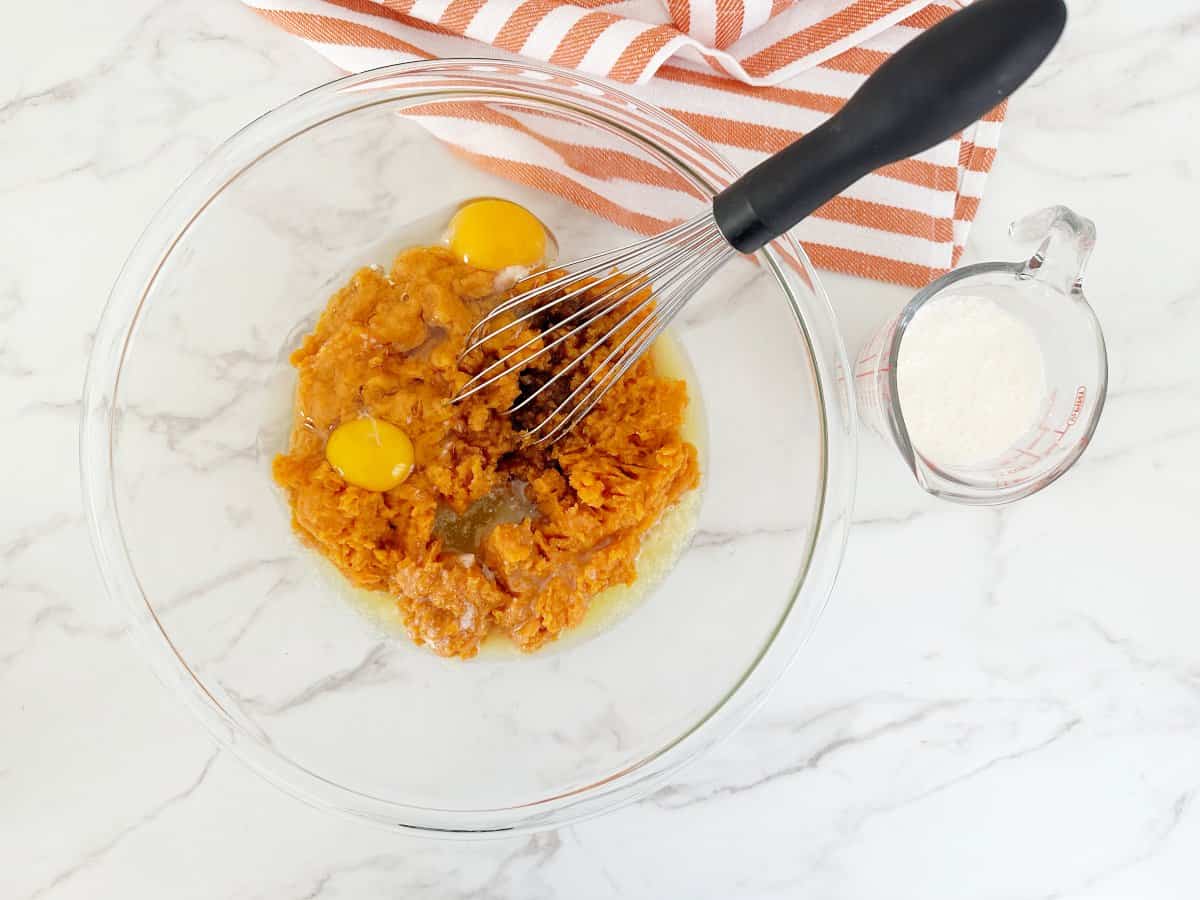 Down view mashed sweet potatoes, eggs, melted butter, vanilla extract in clear bowl with wire whisk. Next to a clear measuring cup with milk.