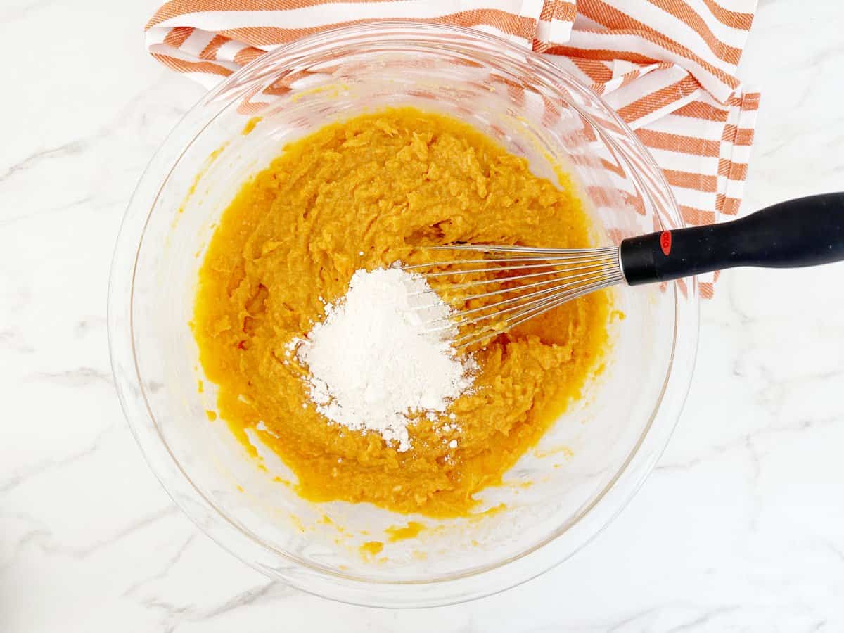 Down view sweet potato mixture and flour in clear bowl with wire whisk.