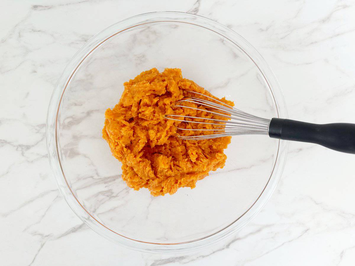 Down view mashed sweet potatoes in clear bowl with wire whisk.