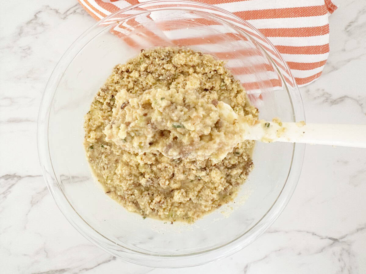 Down view of mixed cornbread stuffing mixture in clear bowl. Hand holding a white spatula showing the very moist mixture.