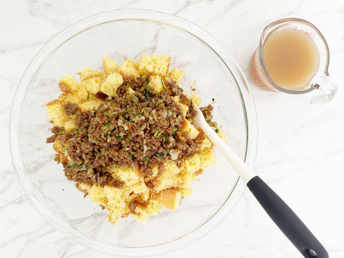 Down view of cubed cornbread and meat vegetable mixture in clear mixing bowl. Next to a glass measuring cup with chicken bone broth.