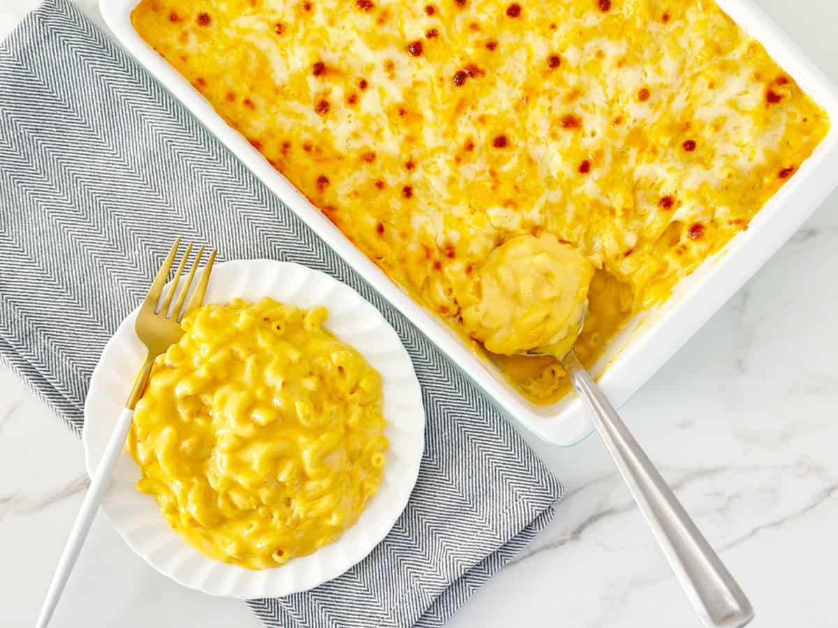 Down view oven baked mac and cheese in baking dish with serving spoon. Next to a white plate with more Mac and cheese with gold fork.