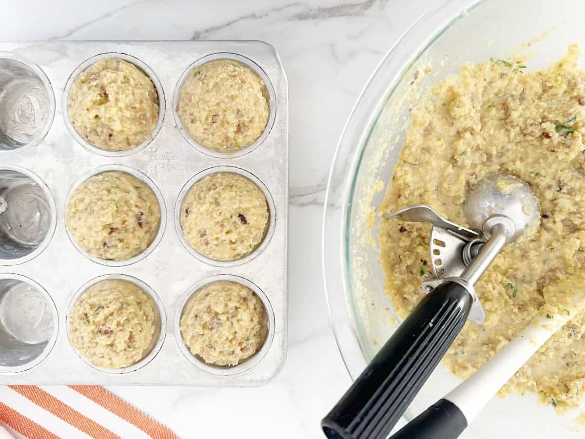 Glass bowl with muffin mixture with ice cream scoop. Next to a muffin tin with portioned stuffing muffins.