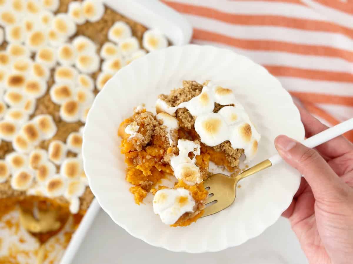 Hand holding white plate and gold fork with a serving of sweet potato casserole.