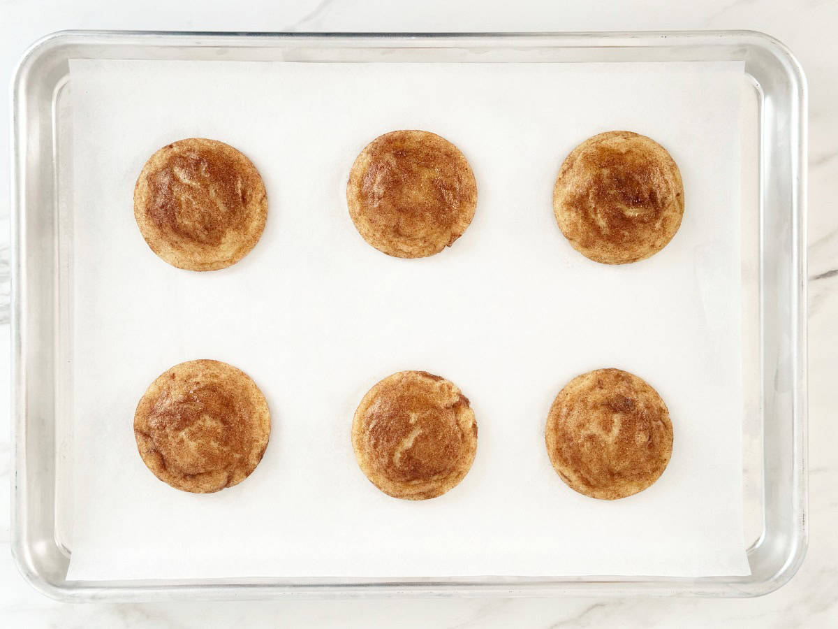 Down view of six baked snickerdoodle cookies on parchment lined baking sheet.