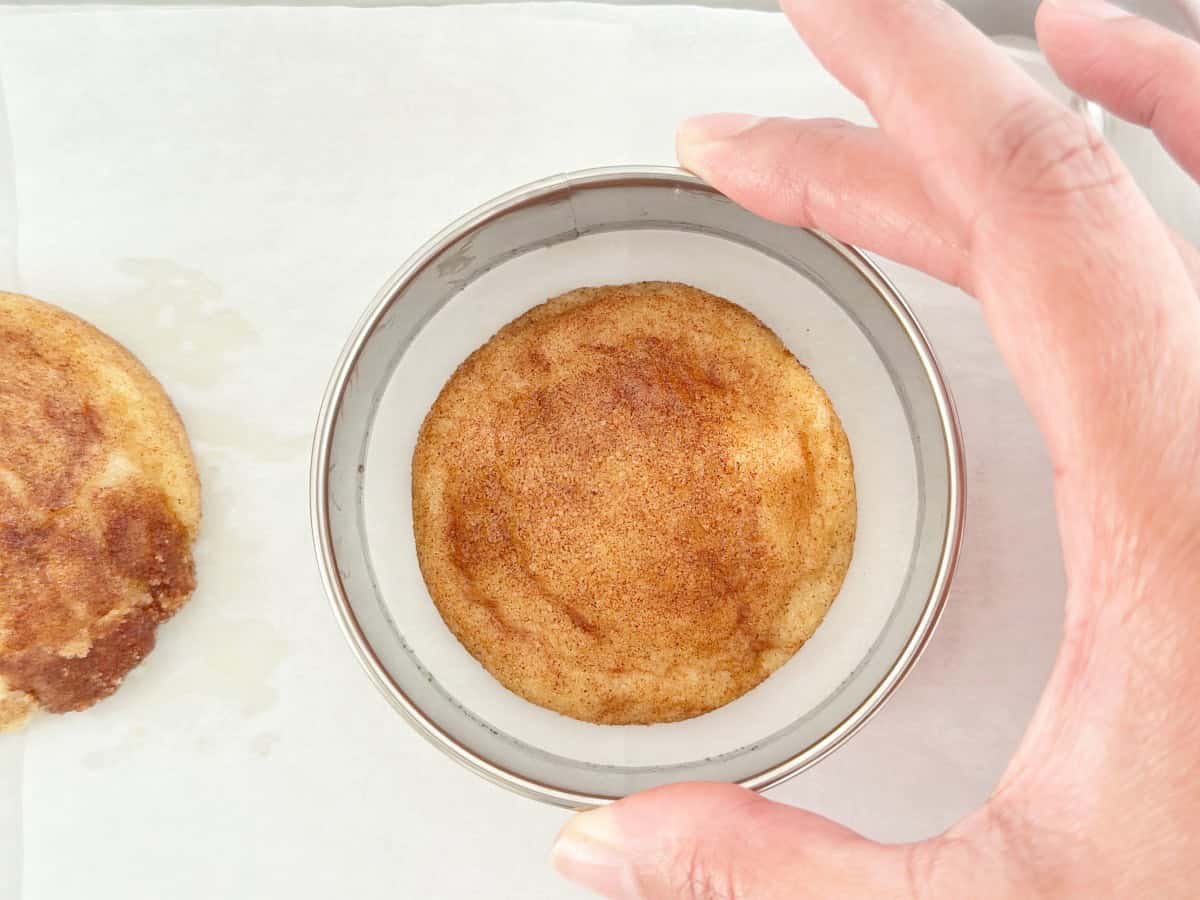 Hand holding a silver round cookie cutter around a snickerdoodle cookie.