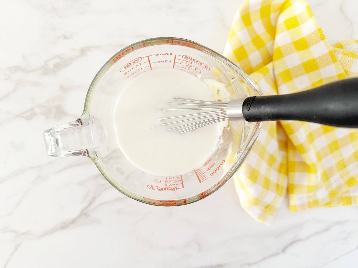 Down view of mixed mayo mixture topped with buttermilk in glass measuring cup with a whisk.