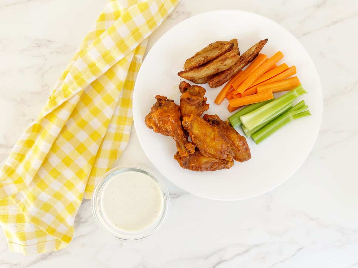 Down view of a white plate with buffalo wings, cut celery and carrots and potato wedges. Next to a glass jar of ranch dressing.