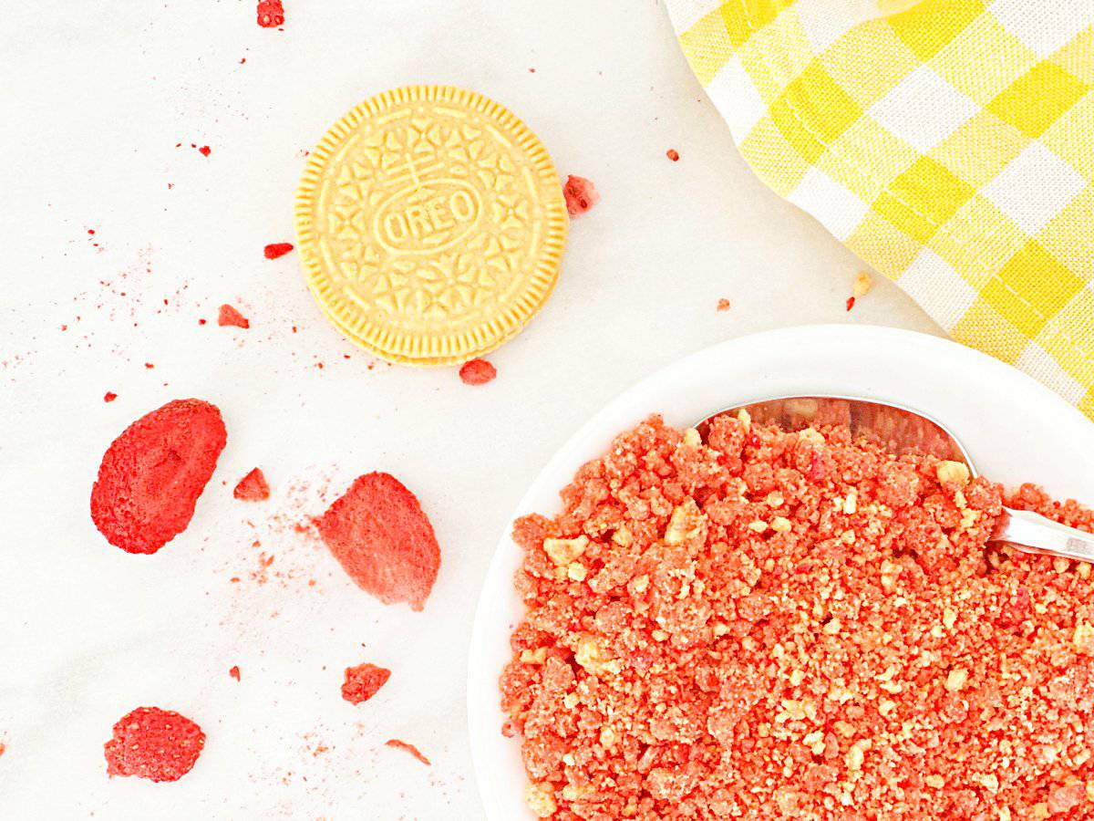 Down view strawberry crunch topping in white bowl with silver spoon. Golden Oreo and freeze dried strawberries next to bowl.