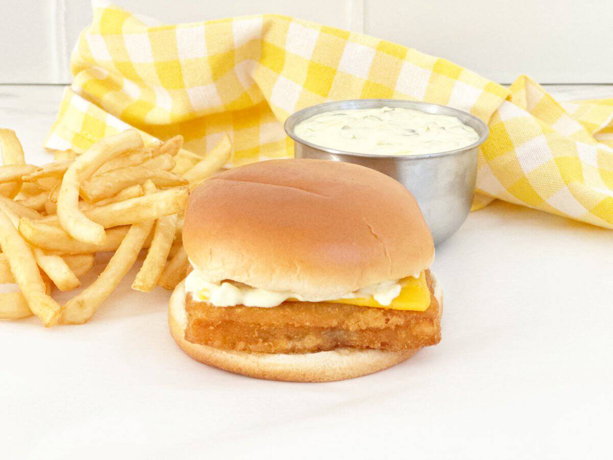 Side view of fish sandwich with tartar sauce in silver bowl and French fries.