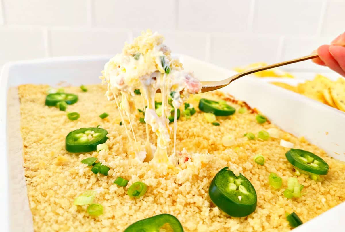 Hand holding a gold spoon with the jalapeรฑo popper chicken dip. The cheesy is stretching from the baking dish.