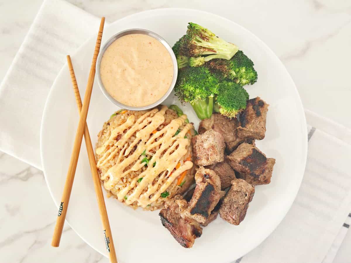 Down view of a white plate with steak hibachi, fried rice, roasted broccoli, silver bowl with yum yum sauce, and chop sticks.