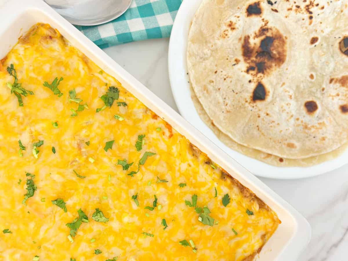 Down view of white baking dish with baked chicken fajita casserole and white plate with two flour tortillas.