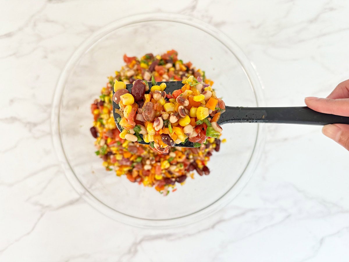 Down view of black rubber spatula holding portion cowboy caviar dip. Under is a mixing bowl with more finished dip.
