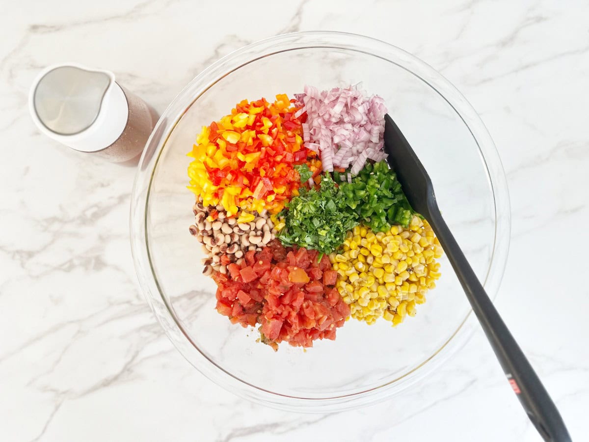 Down view of clear mixing bowl with prepped ingredients and a black rubber spatula. On the side is a salad dressing bottle with taco vinaigrette.