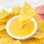 Hand holding tortilla chip with nacho cheese sauce with evaporated milk dripping into white bowl.
