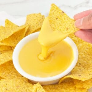 Hand holding tortilla chip with nacho cheese sauce with evaporated milk dripping into white bowl.