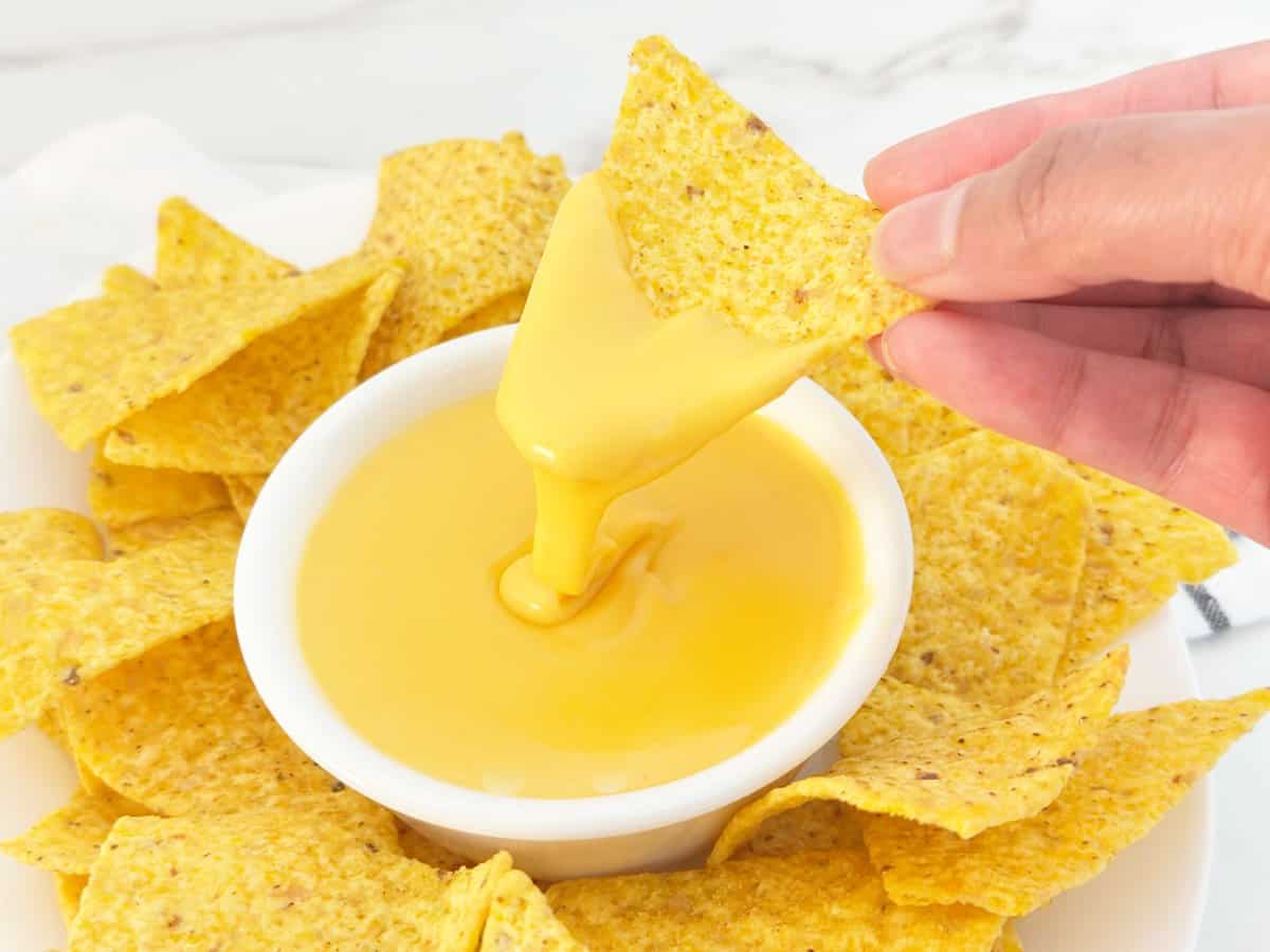 Hand holding tortilla chip with nacho cheese sauce with evaporated milk dripping into white bowl.