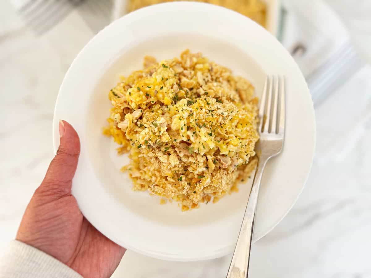 Hand holding a white plate with a serving portion of old fashioned chicken and rice casserole and a fork.