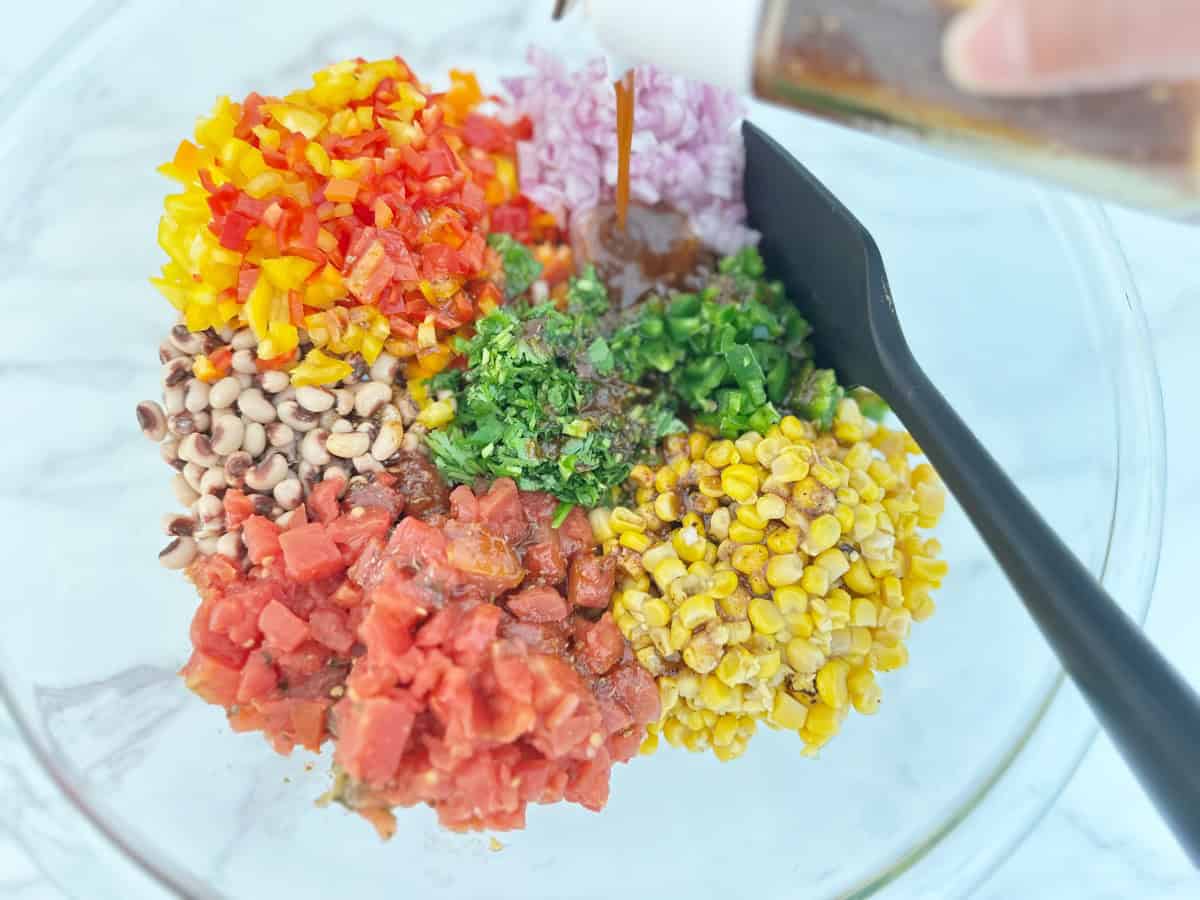 Down view of clear mixing bowl with prepped ingredients and a black rubber spatula. Hand pouring dressing into mixing bowl.