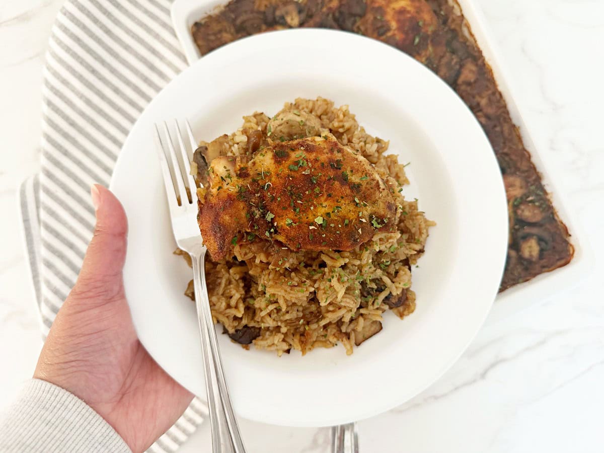 Hand holding a white plate with a serving portion of stick of butter rice with chicken and a fork.