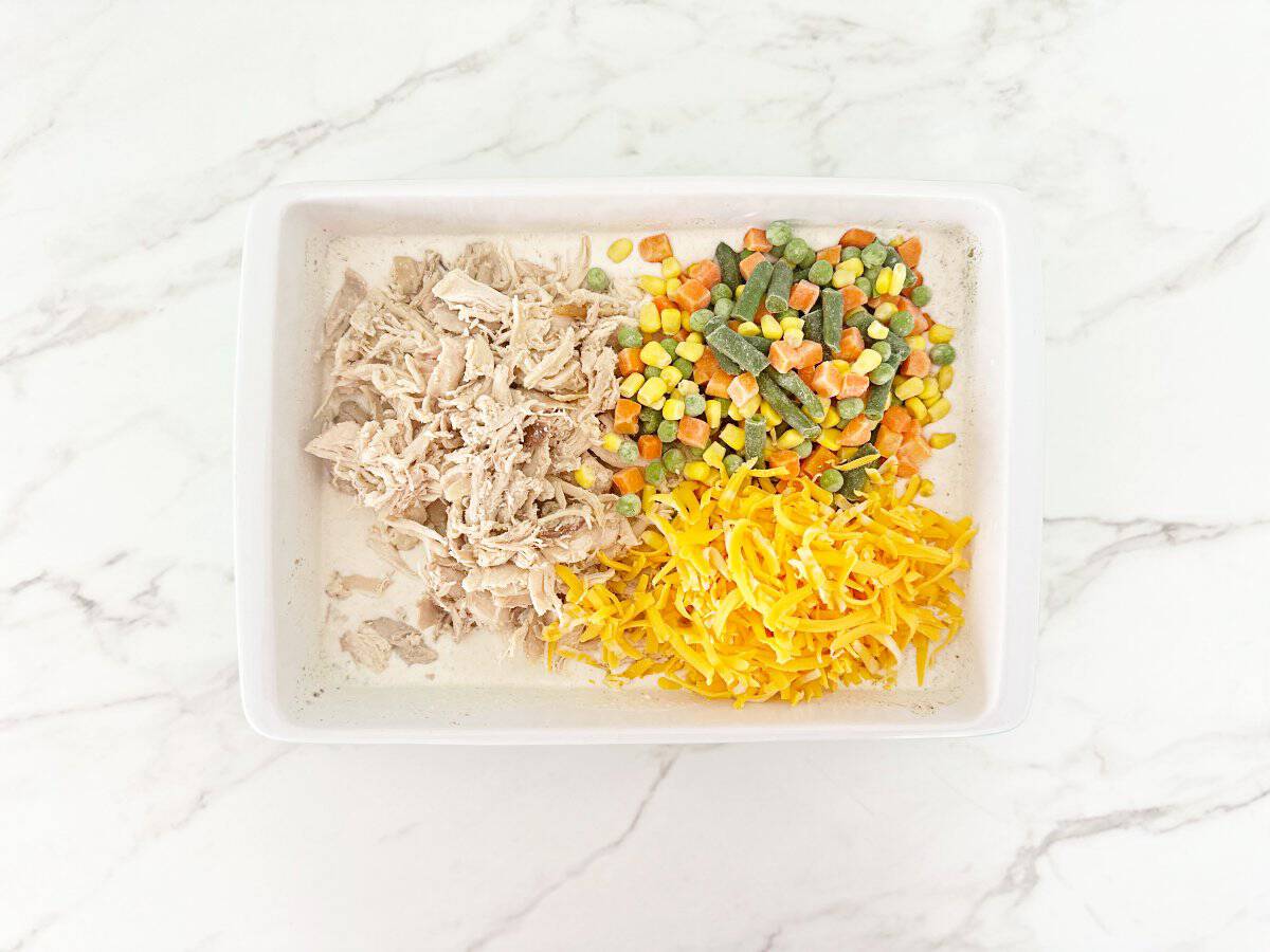 Down view white baking dish with cooked chicken, shredded cheese, and frozen mixed vegetables.