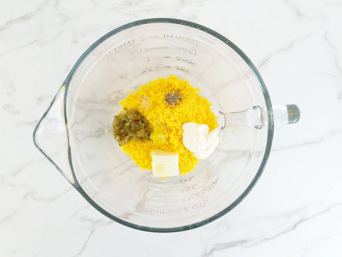Down view of a glass mixing bowl with smooth egg yolks, butter, relish, pepper, and mayo.
