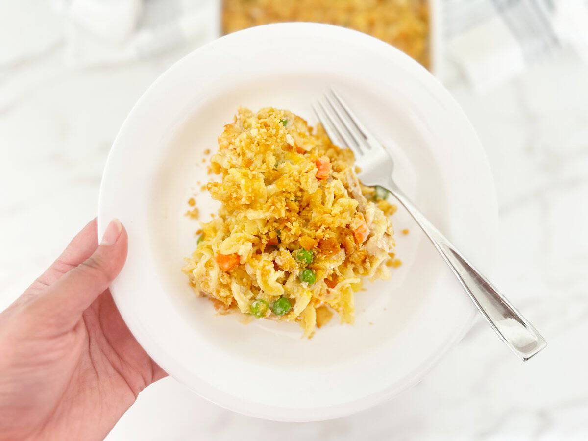 Hand holding a white plate with a serving portion of old fashioned chicken noodle casserole and a fork.