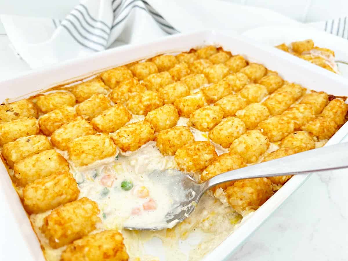 Side view of creamy chicken pot pie with tater tots in white baking dish. Shows creamy texture on silver serving spoon.