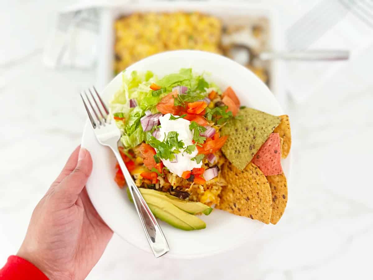 Hand holding a chicken and yellow rice bowl topped with sour cream, tomatoes and cilantro. Side of avocado and tortilla chips.