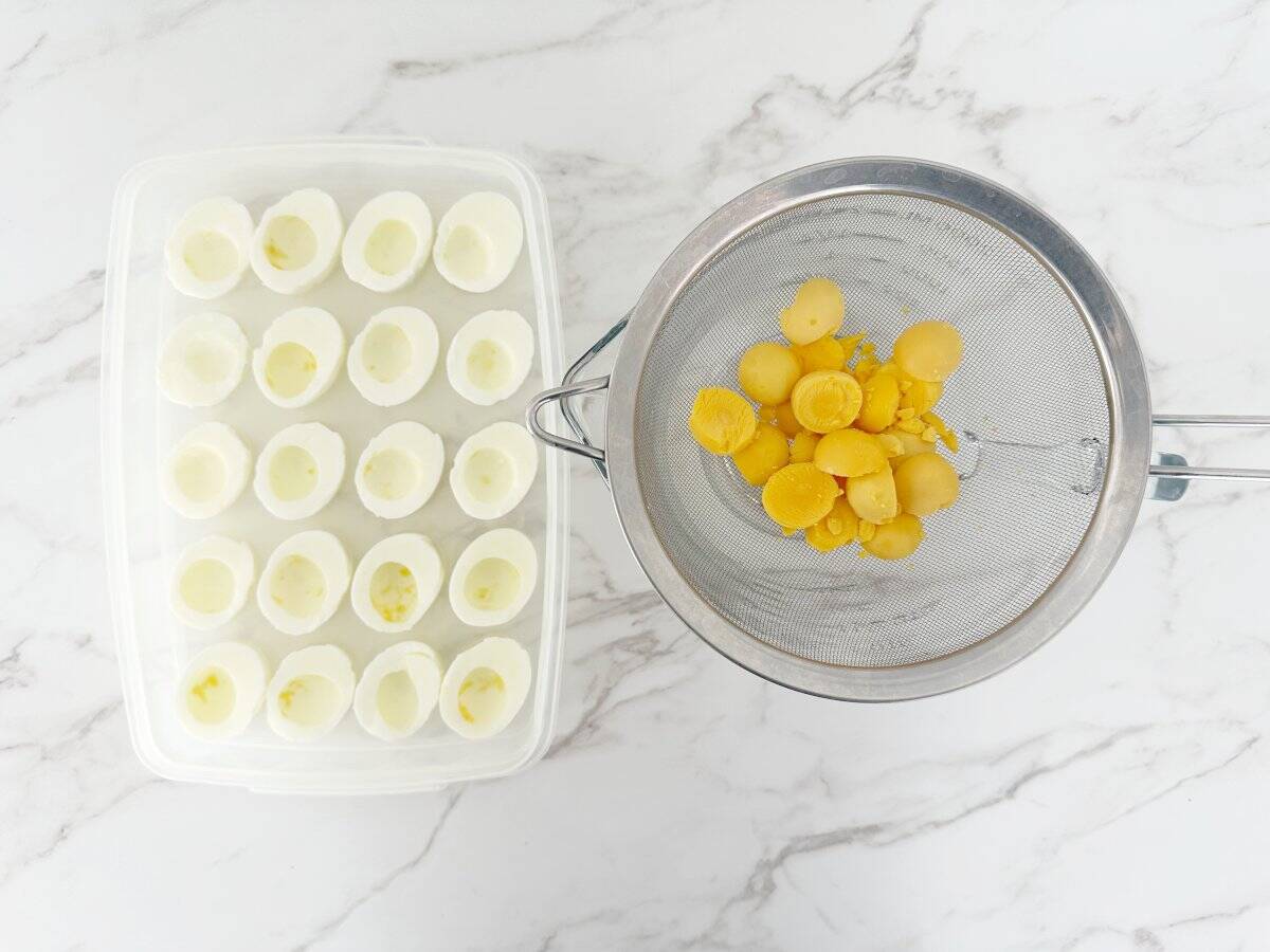 Down view of a plastic deviled egg container with cooked egg whites. Next to a strainer with egg yolks.