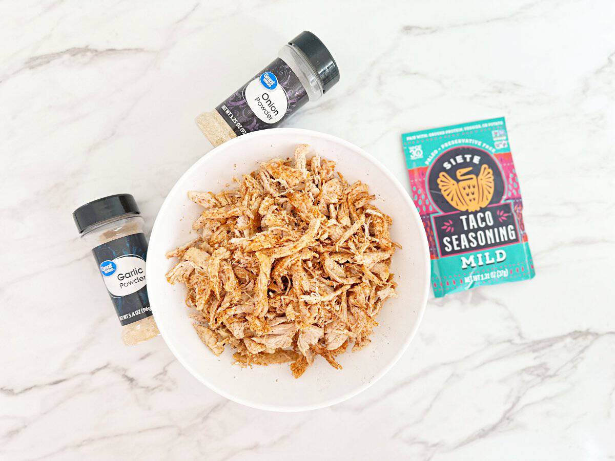 Down view of white bowl with taco seasoned shredded rotisserie chicken. Next to a packet of taco seasoning, bottle onion powder and bottle of garlic powder.