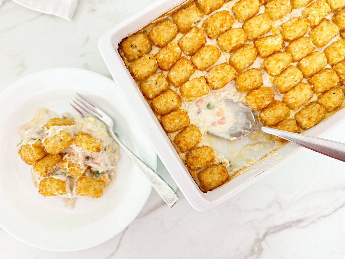 Down view of tater tot chicken pot pie casserole in white baking dish. Next to a dinner serving of the casserole in white plate and fork.