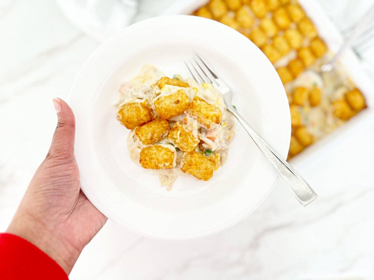 Hand holding a white plate with a serving portion of tater tot chicken pot pie casserole and a fork.
