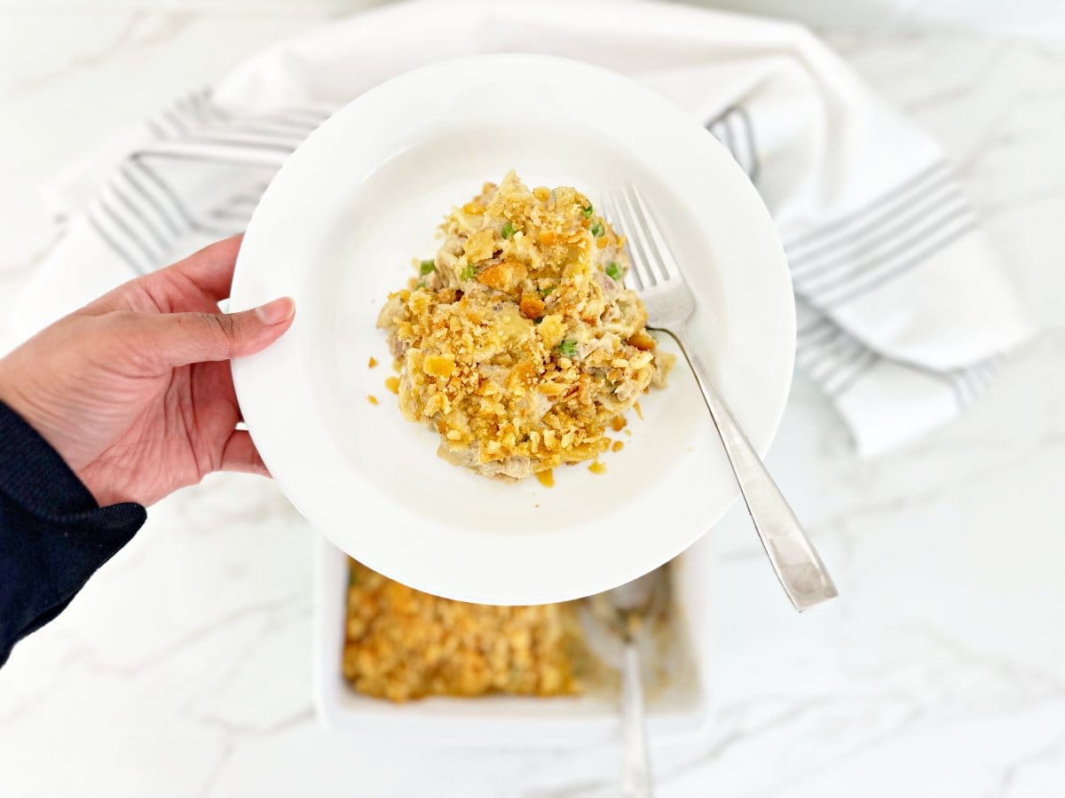 Hand holding a white plate with a serving portion of tuna noodle casserole and a fork.