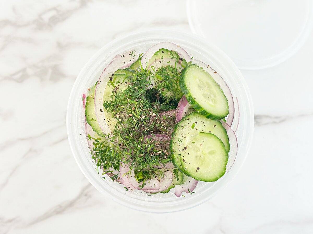 Down view of deli container with cucumber, onions, spices, and fresh dill.