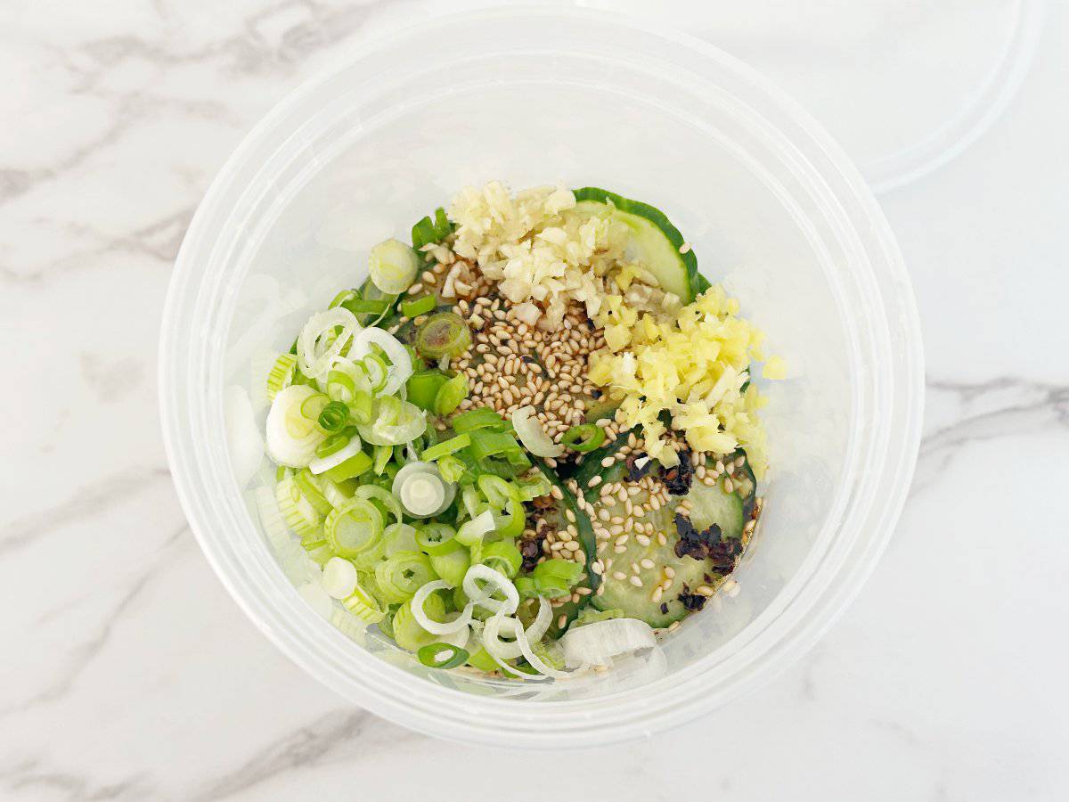 Down view of deli container with cucumber, rice vinegar, soy sauce, green onion, garlic, ginger, and sesame seeds.