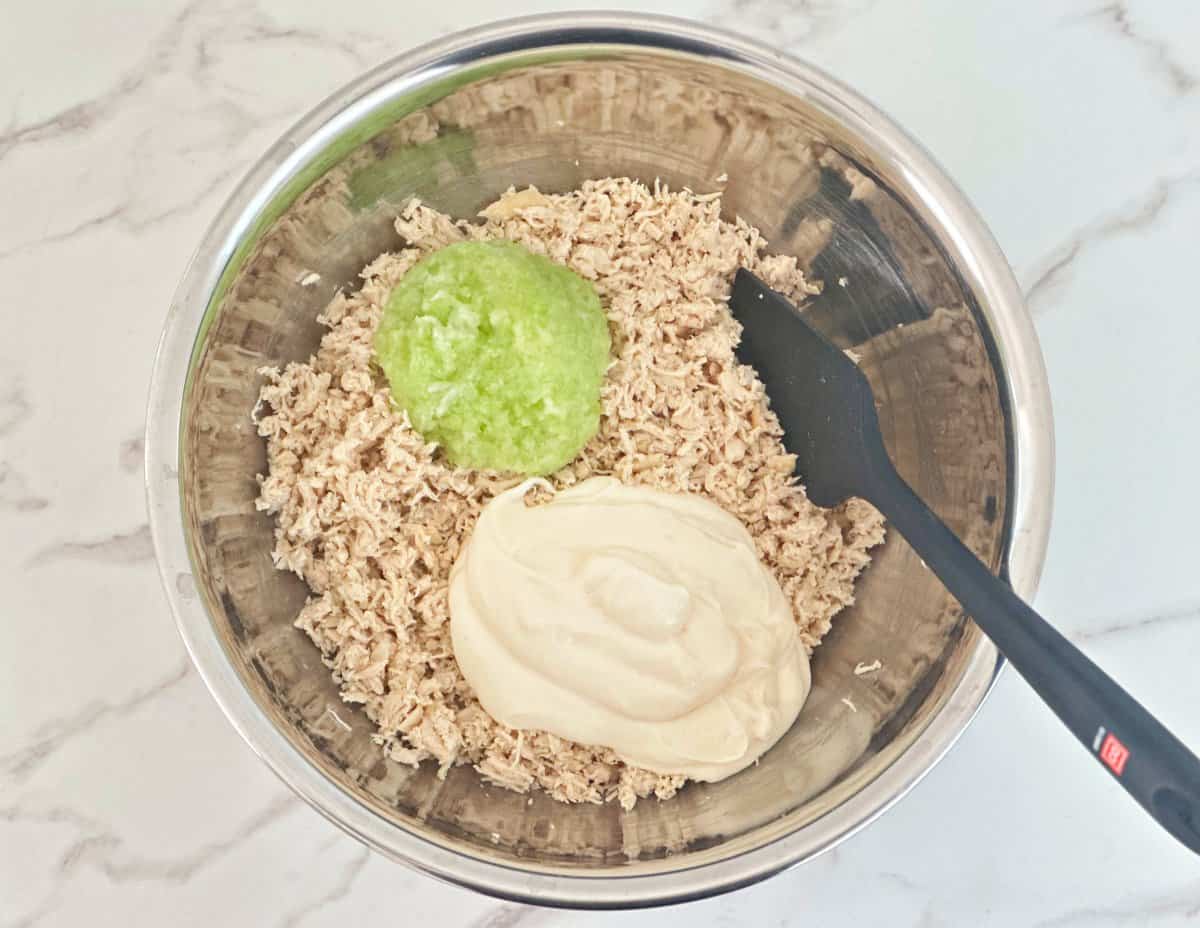 Down view of silver mixing bowl with shredded chicken, celery, onion, and mayo.
