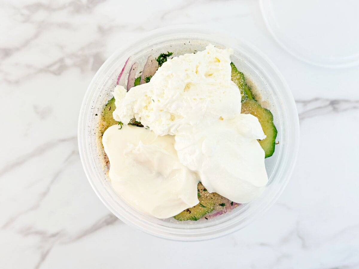 Down view of deli container with cucumber, onions, spices, dill, mayo, sour cream and cream cheese.