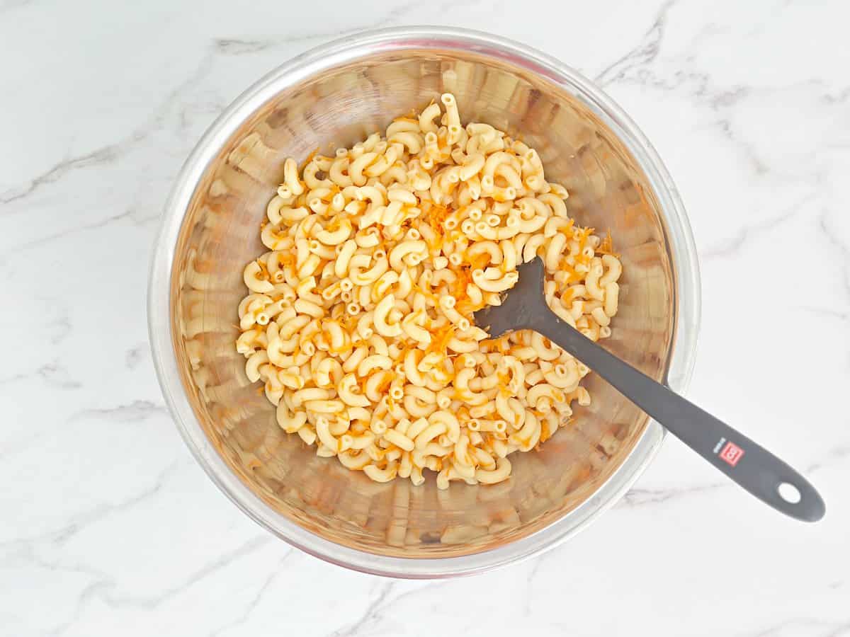 Down view silver mixing bowl with cooked macaroni, onion, vinegar, and shredded carrot.