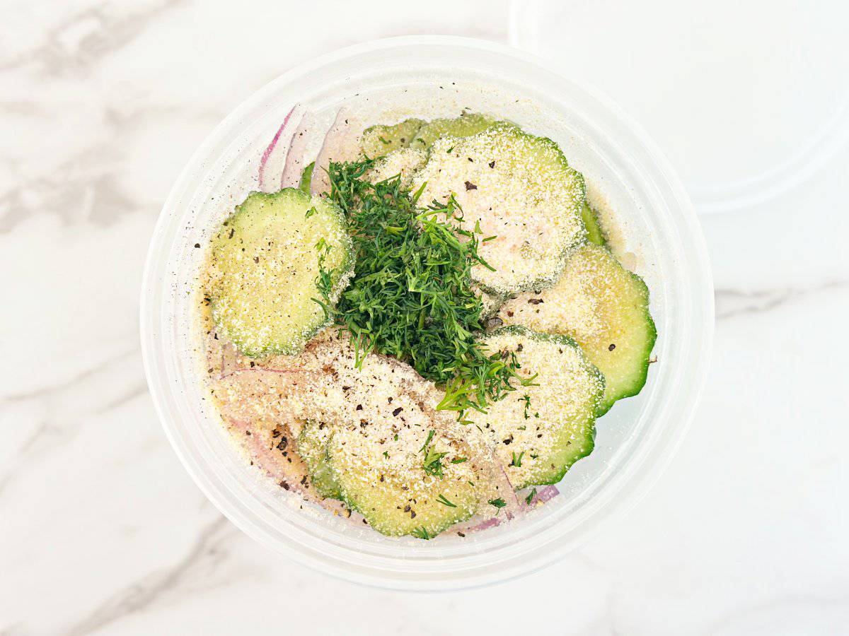 Down view of deli container with cucumber, onions, spices, and fresh dill.