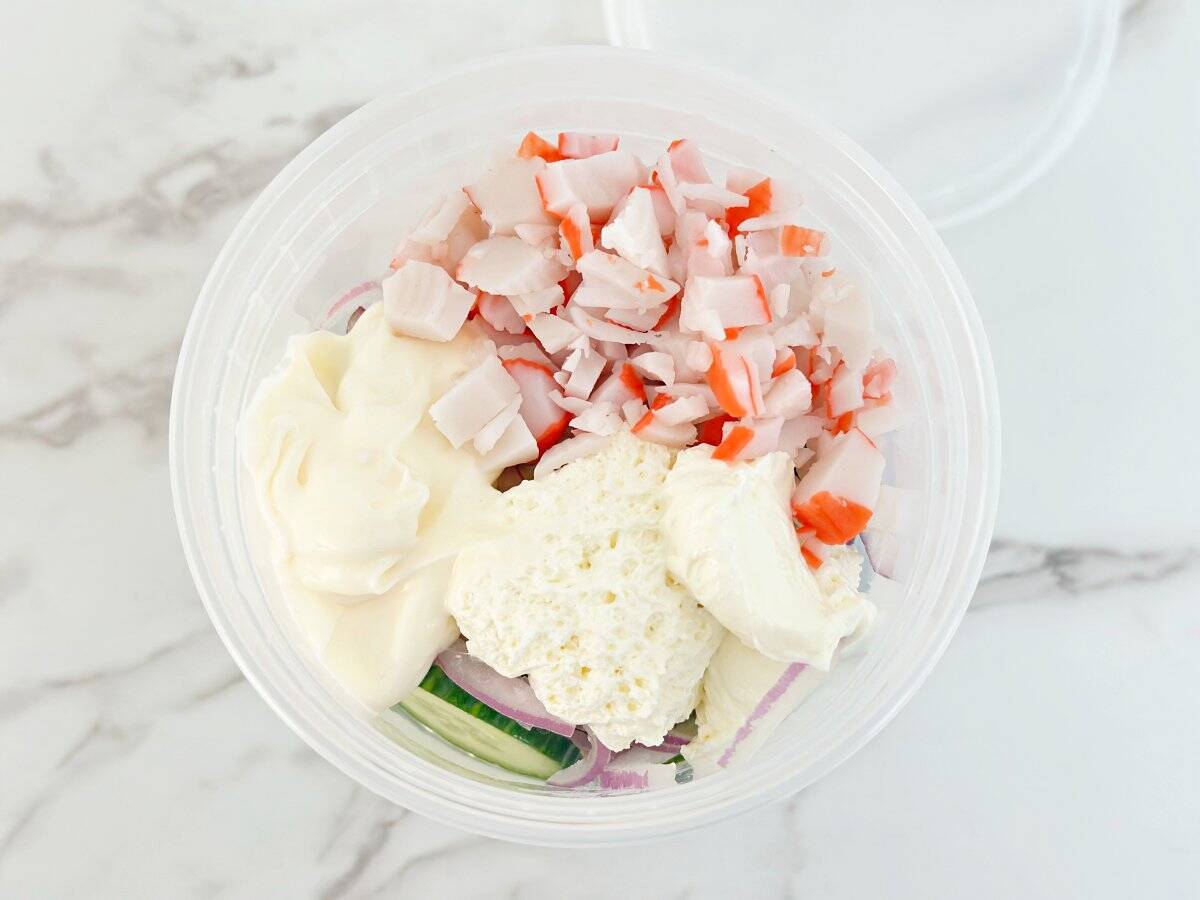 Down view of deli container with cucumber, onions, spices, mayo, chopped imitation crab, and cream cheese.