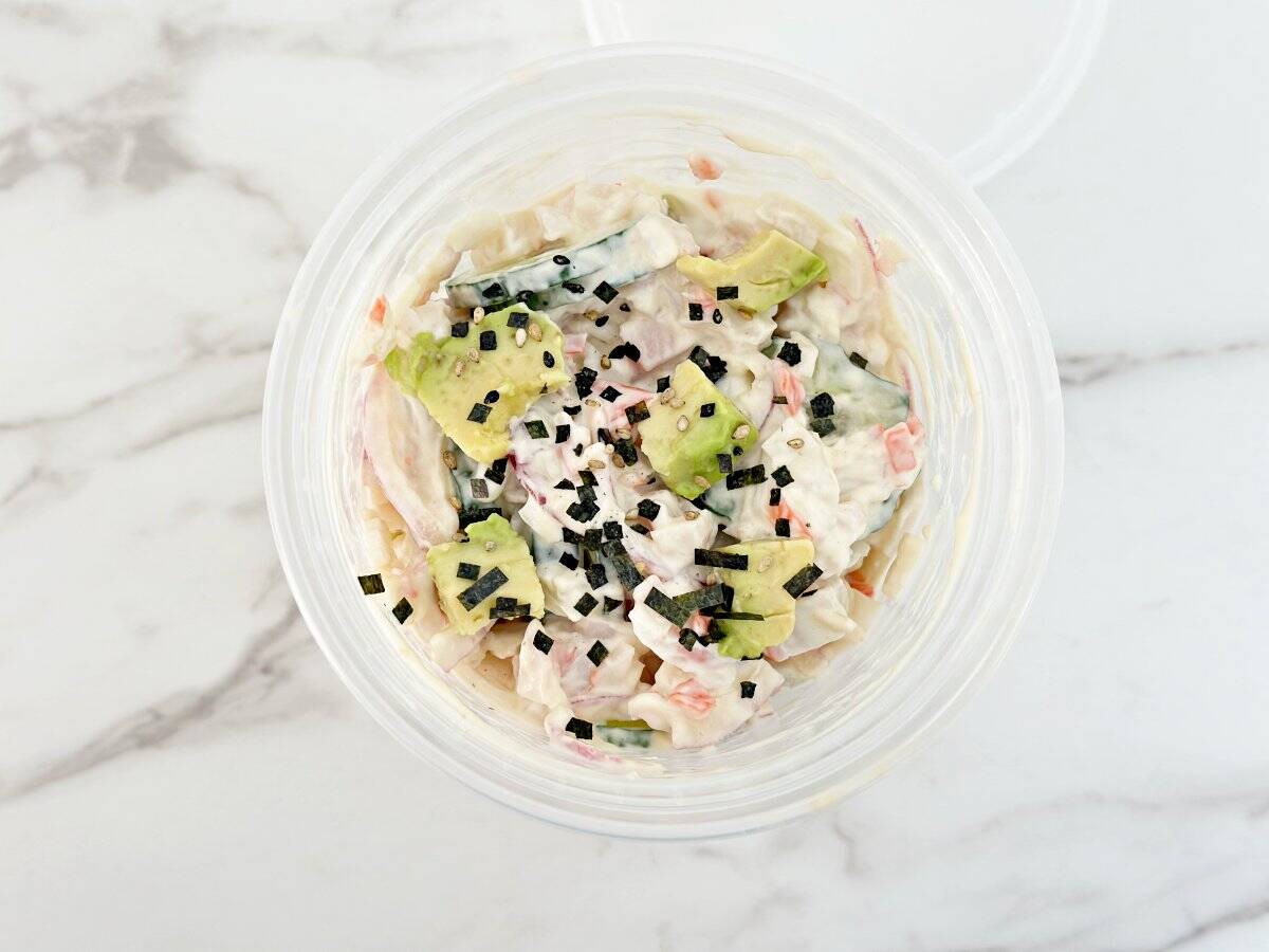 Down view of deli container with sushi cumber salad topped with avocado and furikaki.
