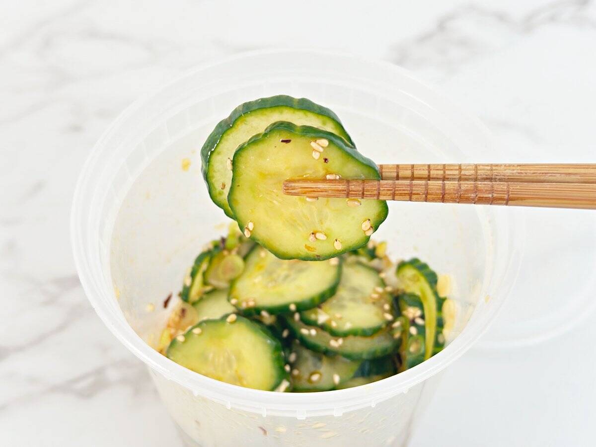 Chopstick holding a portion of asian cucumber salad from a deli container.