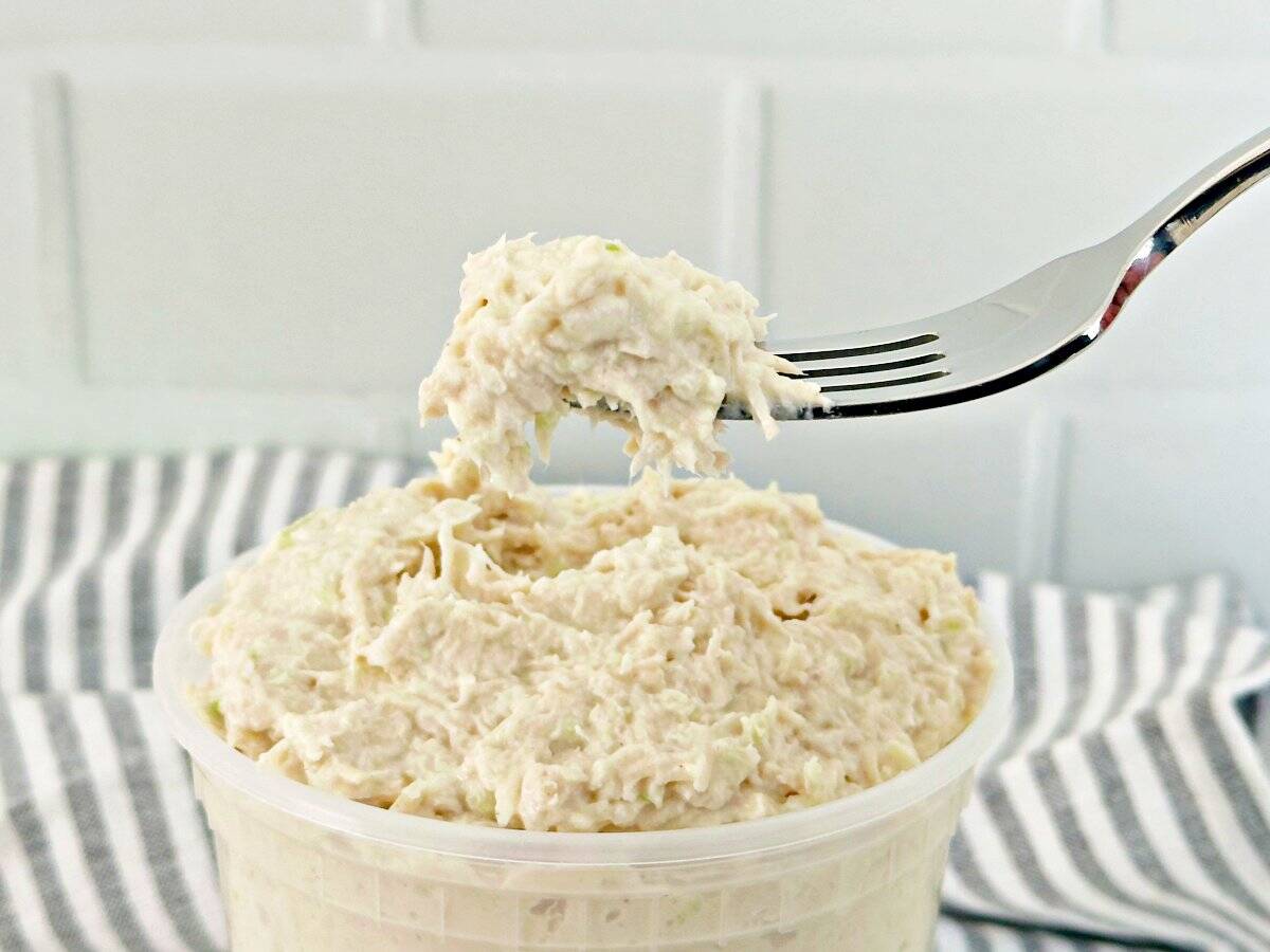 A small portion of chicken salad on a silver fork. Under is a deli container with creamy chicken salad.