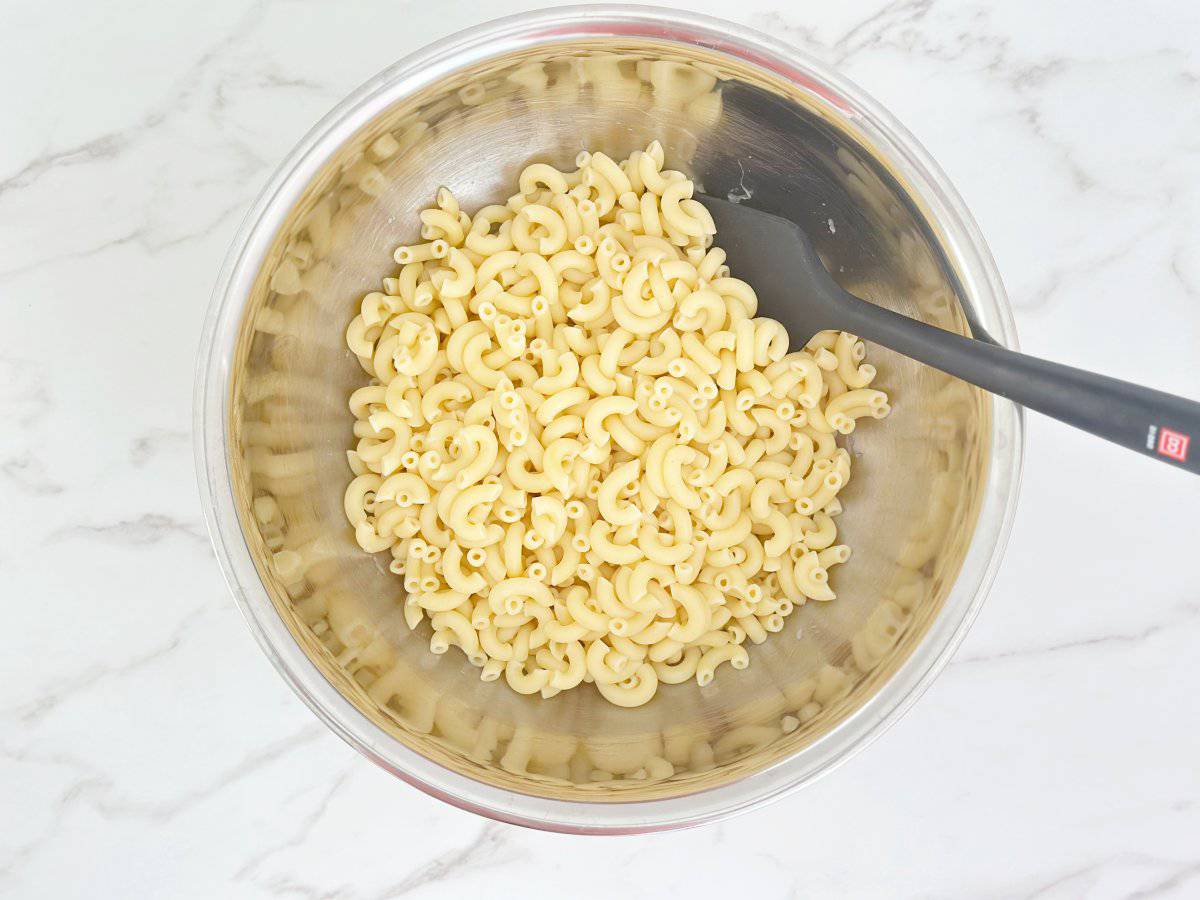 Down view silver mixing bowl with cooked macaroni, onion, vinegar, and sugar.