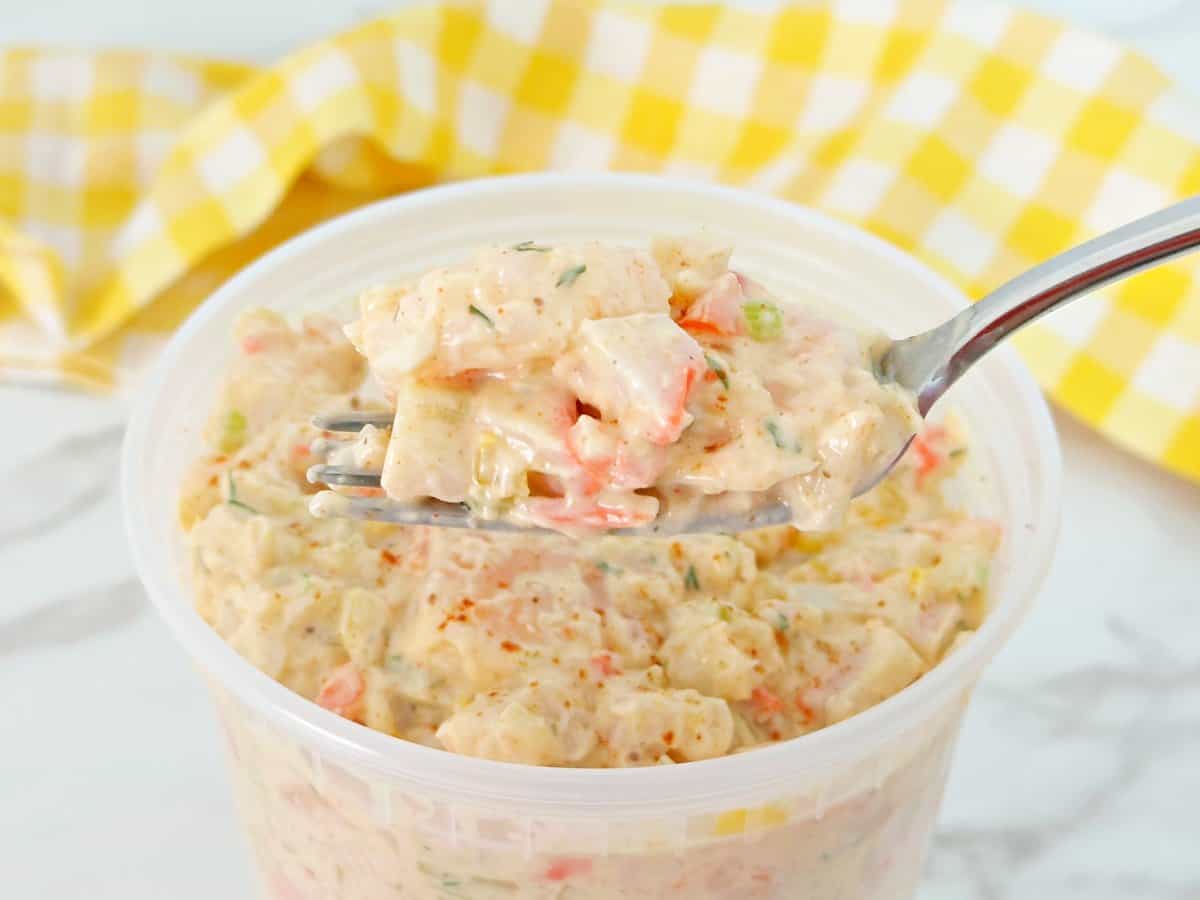 A small portion of seafood salad on a silver fork. Under is a deli container with creamy seafood salad.