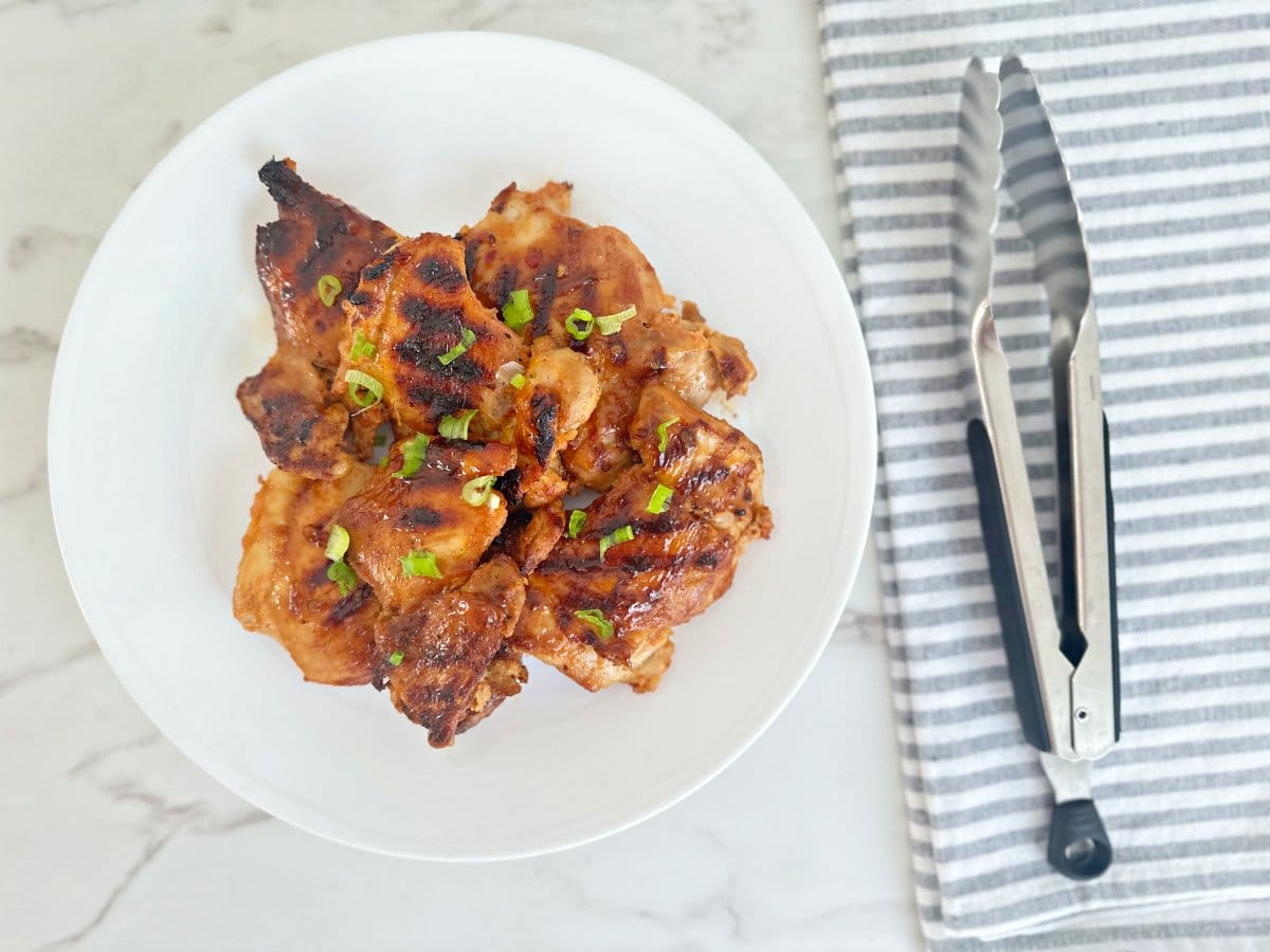 Down view of huli huli Hawaiian chicken thighs on white plate next to a grilling tongs.