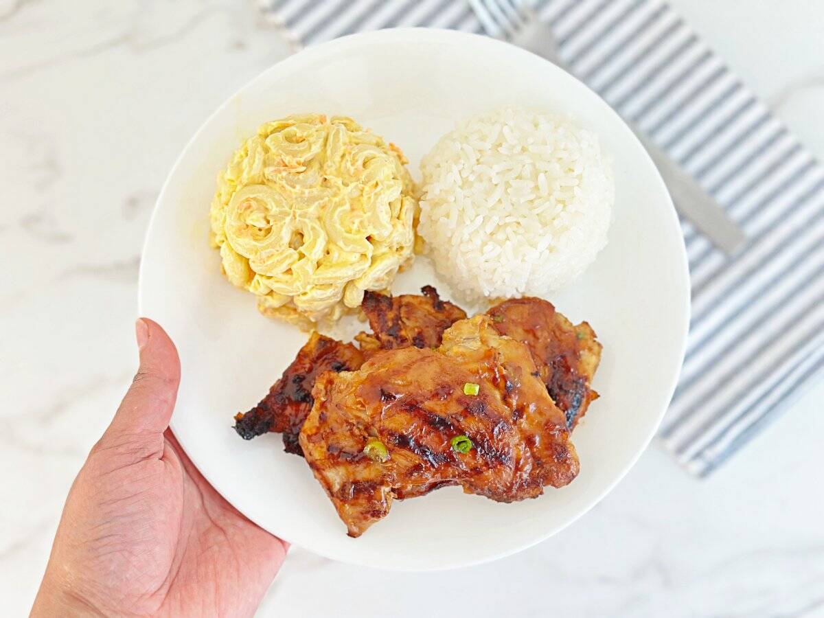 Hand holding a white plate of huli huli Hawaiian chicken bbq. Side of Macaroni salad, white rice, and grilled chicken thighs.
